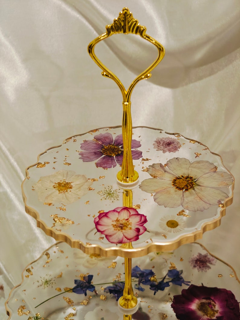 K&ouml;nnte beinhalten: Ein zweist&ouml;ckiger Kuchenst&auml;nder mit einem goldenen Metallrahmen und Griff. Die obere Etage hat einen wei&szlig;en Hintergrund mit goldenen Blattverzierungen und gepressten Blumen in Violett-, Wei&szlig;- und Rosat&ouml;nen. Die untere Etage hat ein &auml;hnliches Design mit blauen und violetten Blumen.
