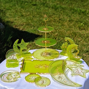 May include: A collection of green and gold resin serving pieces and decorative items. Includes a three-tiered stand, trays, coasters, and star and crescent moon shapes. The items are arranged on a white tablecloth outdoors.