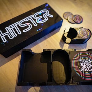 Op de afbeelding: Een zwarte doos en dienblad set met het woord "HITSTER" in het wit. De doos heeft een sterrenpatroon. Een houder met het woord "HITSTER" en verschillende ronde tokens zijn ook zichtbaar. Een kaart met een QR-code staat in de lade.