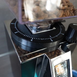 Op de afbeelding: Close-up van een moderne koffiemolen met een doorzichtige bonencontainer gevuld met geroosterde koffiebonen. De molen heeft een zwarte draaiknop met witte markeringen en een digitaal display. De machine is gemaakt van roestvrij staal.