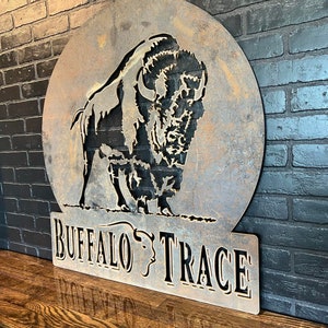 Puede incluir: Un letrero de metal oxidado con una silueta de b&uacute;falo en un c&iacute;rculo y el texto "Buffalo Trace" debajo.