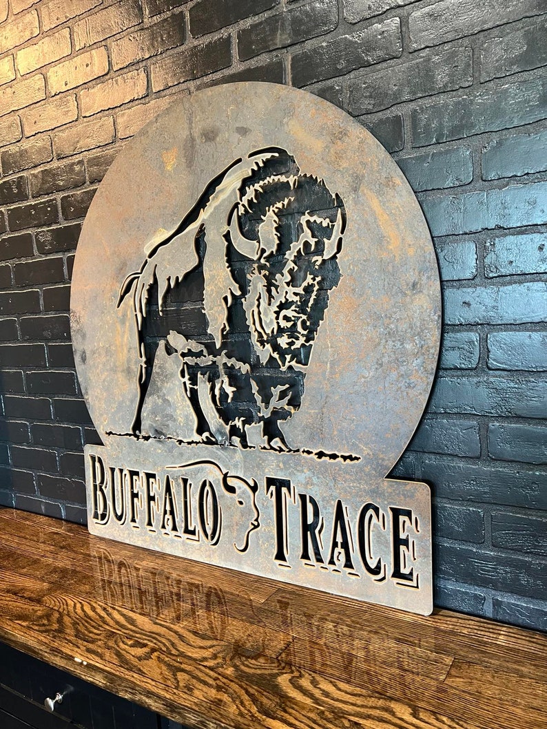Puede incluir: Un letrero de metal oxidado con una silueta de b&uacute;falo en un c&iacute;rculo y el texto "Buffalo Trace" debajo.