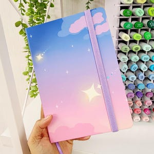 May include: A notebook with a dreamy sky design. The cover transitions from blue to pink, featuring clouds, stars, and a shooting star. A lavender elastic band secures the notebook. The notebook is held by a person.
