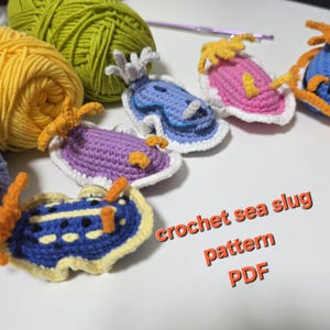 Może przedstawiać: Kolekcja kolorowych wzorów na szydełkowe ślimaki morskie w różnych kolorach, w tym niebieskim, fioletowym, różowym i żółtym. Obraz przedstawia również kłęby żółtej i zielonej przędzy oraz tekst "crochet sea slug pattern PDF".