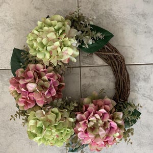 May include: A circular wreath with a base made of interwoven twigs, decorated with pink and green hydrangea flowers. The wreath is embellished with green leaves and small sprigs of foliage, creating a natural, colourful floral display.
