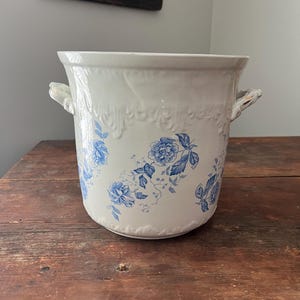 May include: A white ceramic planter with blue floral designs and two small handles. The planter has a decorative embossed pattern around the top. The floral design features blue flowers and leaves. The planter is on a wooden surface.