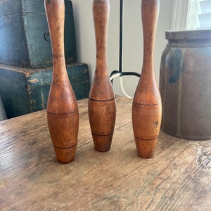 May include: Three tall, slender wooden bowling pins with a weathered, medium-brown finish. Each pin features a rounded top, a tapered neck, and a wider base, accented by two thin, horizontal black rings encircling the lower portion of the body.