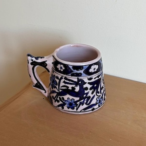 May include: A ceramic mug with a pink exterior and interior. The mug features a blue and white floral and animal design. The handle is pink with black accents. The mug sits on a wooden surface.