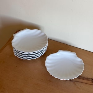 May include: A set of seven white, scallop-shaped plates. Six plates are stacked, with a seventh plate placed nearby. Each plate features a wavy, textured edge and a smooth, glossy finish. The plates are on a wooden surface.