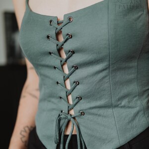 May include: A sage green sleeveless corset-style top featuring a front lace-up closure with dark metal eyelets and matching fabric ties. The garment has a straight neckline, thin shoulder straps, and vertical seam detailing along the front panels.