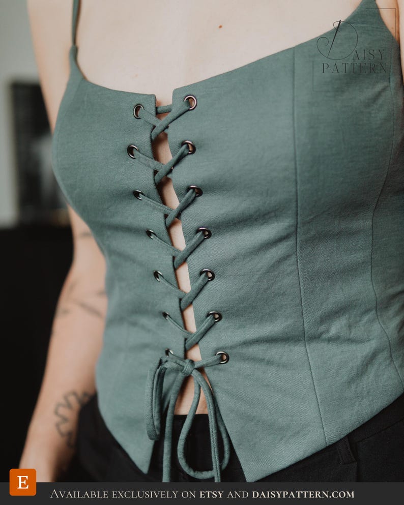 May include: A sage green sleeveless corset-style top featuring a front lace-up closure with dark metal eyelets and matching fabric ties. The garment has a straight neckline, thin shoulder straps, and vertical seam detailing along the front panels.