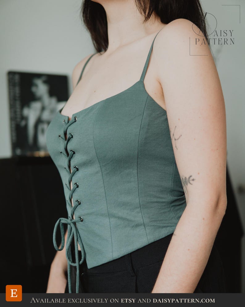May include: A sage green corset-style top featuring thin spaghetti straps, a front lace-up closure with silver-toned metal eyelets, and a tie detail at the hem. The garment is constructed with vertical seams and has a fitted silhouette.