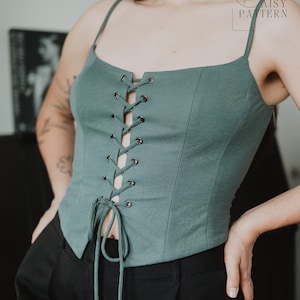 May include: A sage green corset-style top featuring thin spaghetti straps, a square neckline, and a front lace-up closure with silver-toned metal eyelets and matching fabric ties. The garment has a fitted silhouette with vertical seam detailing.