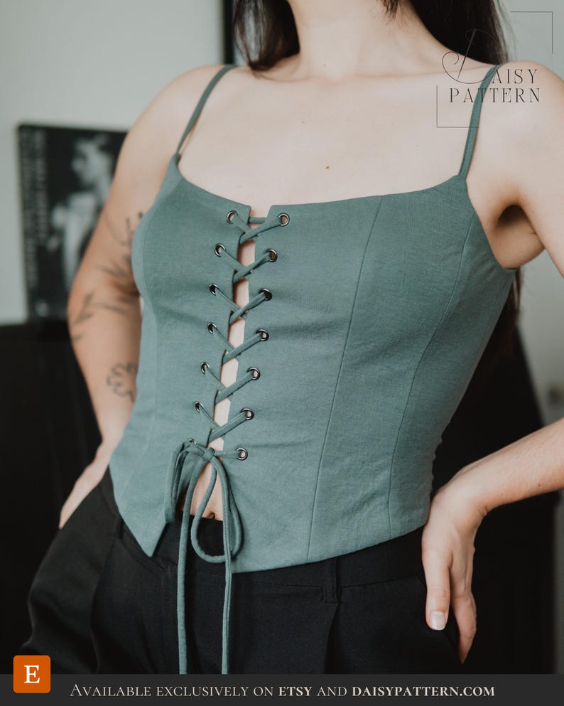 May include: A sage green corset-style top featuring thin spaghetti straps, a square neckline, and a front lace-up closure with silver-toned metal eyelets and matching fabric ties. The garment has a fitted silhouette with vertical seam detailing.