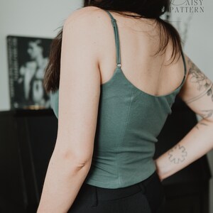 May include: A sage green camisole top featuring thin, adjustable spaghetti straps and a scoop neckline. The garment is constructed with vertical seam detailing along the back and sides, providing a fitted silhouette. The fabric appears to be a solid, matte-finish jersey material.