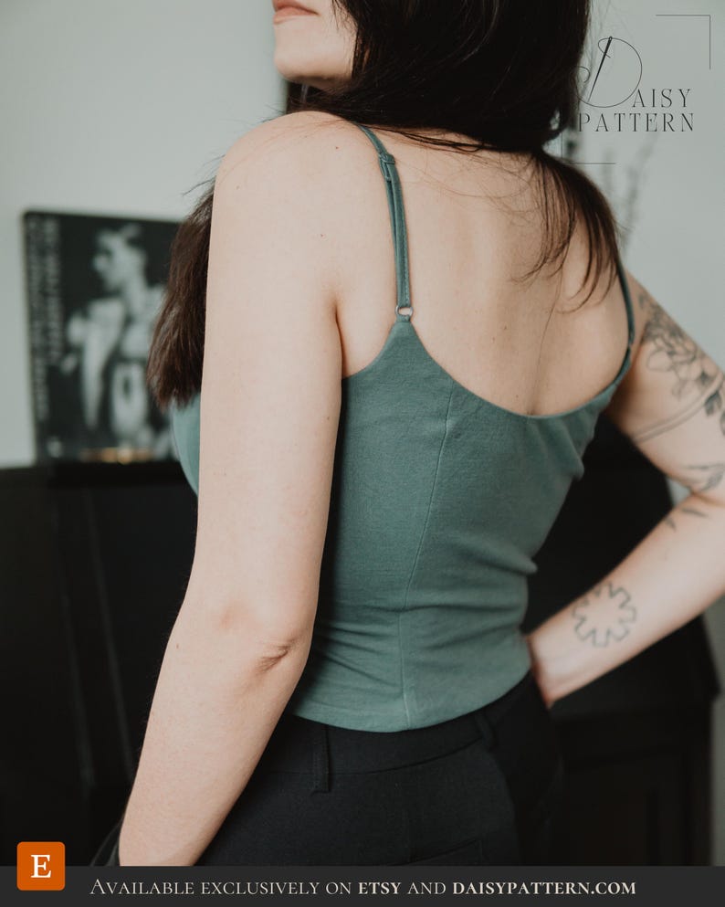May include: A sage green camisole top featuring thin, adjustable spaghetti straps and a scoop neckline. The garment is constructed with vertical seam detailing along the back and sides, providing a fitted silhouette. The fabric appears to be a solid, matte-finish jersey material.