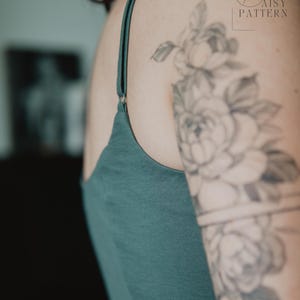 May include: A dark teal tank top featuring thin adjustable shoulder straps with metal sliders. The garment has a simple neckline and is shown from a side angle, revealing the fabric texture and the solid, deep green color of the material.