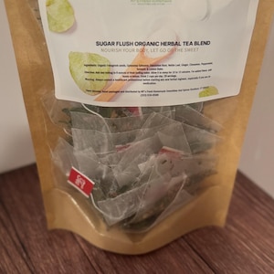 Puede incluir: Una bolsa de papel marrón con una etiqueta blanca que dice "SUGAR FLUSH ORGANIC HERBAL TEA BLEND NOURISH YOUR BODY, LET GO OF THE SWEET". La bolsa contiene una variedad de bolsitas de té de hojas sueltas.