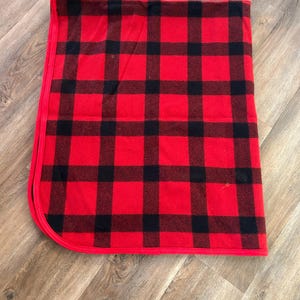 Może przedstawiać: Złożony czerwono-czarny koc w kratę. Koc ma klasyczny wz&oacute;r w kratę z odważnymi czerwonymi i czarnymi kwadratami. Krawędzie są starannie wykończone czerwoną obw&oacute;dką, tworząc przytulną i rustykalną estetykę.