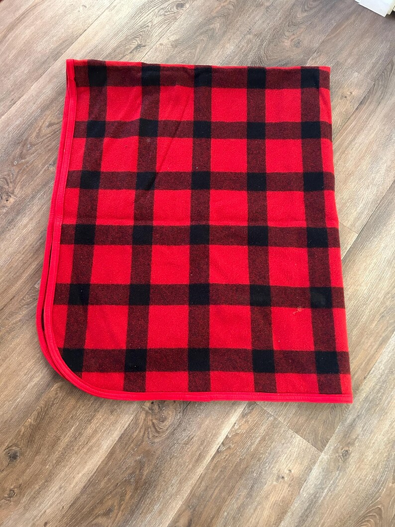 Może przedstawiać: Złożony czerwono-czarny koc w kratę. Koc ma klasyczny wz&oacute;r w kratę z odważnymi czerwonymi i czarnymi kwadratami. Krawędzie są starannie wykończone czerwoną obw&oacute;dką, tworząc przytulną i rustykalną estetykę.