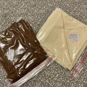 May include: Two fabric items, one brown and one cream, are sealed in clear plastic bags with red zipper closures. The brown fabric is folded and appears shiny. The cream fabric is folded neatly with a label attached. The items are on a patterned surface.