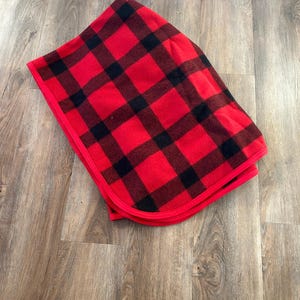 Puede incluir: Una manta plegada a cuadros rojos y negros. La manta presenta un patrón de cuadros clásico con grandes cuadrados negros sobre un fondo rojo. Los bordes están acabados con un borde rojo. La manta está doblada cuidadosamente.