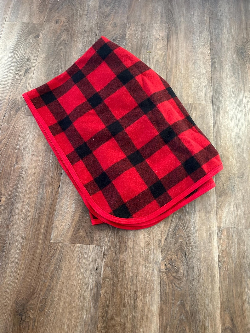 Może przedstawiać: Złożony czerwono-czarny koc w kratę. Koc ma klasyczny wz&oacute;r w kratę z dużymi czarnymi kwadratami na czerwonym tle. Krawędzie wykończone są czerwoną obw&oacute;dką. Koc jest starannie złożony.