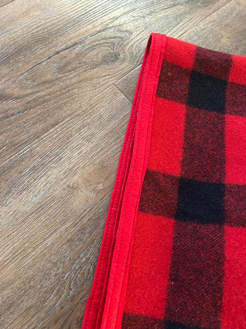 Może przedstawiać: Złożony czerwono-czarny koc w kratę. Koc ma klasyczny wz&oacute;r w kratę w odważnych czerwonych i czarnych kwadratach. Krawędź koca jest obszyta jednolitym czerwonym obramowaniem.