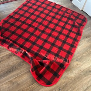 Może przedstawiać: Czerwono-czarny koc w kratę z zaokrągloną krawędzią. Koc jest wykonany z miękkiego materiału i jest złożony na drewnianej podłodze. Wz&oacute;r to klasyczna krata bawola.