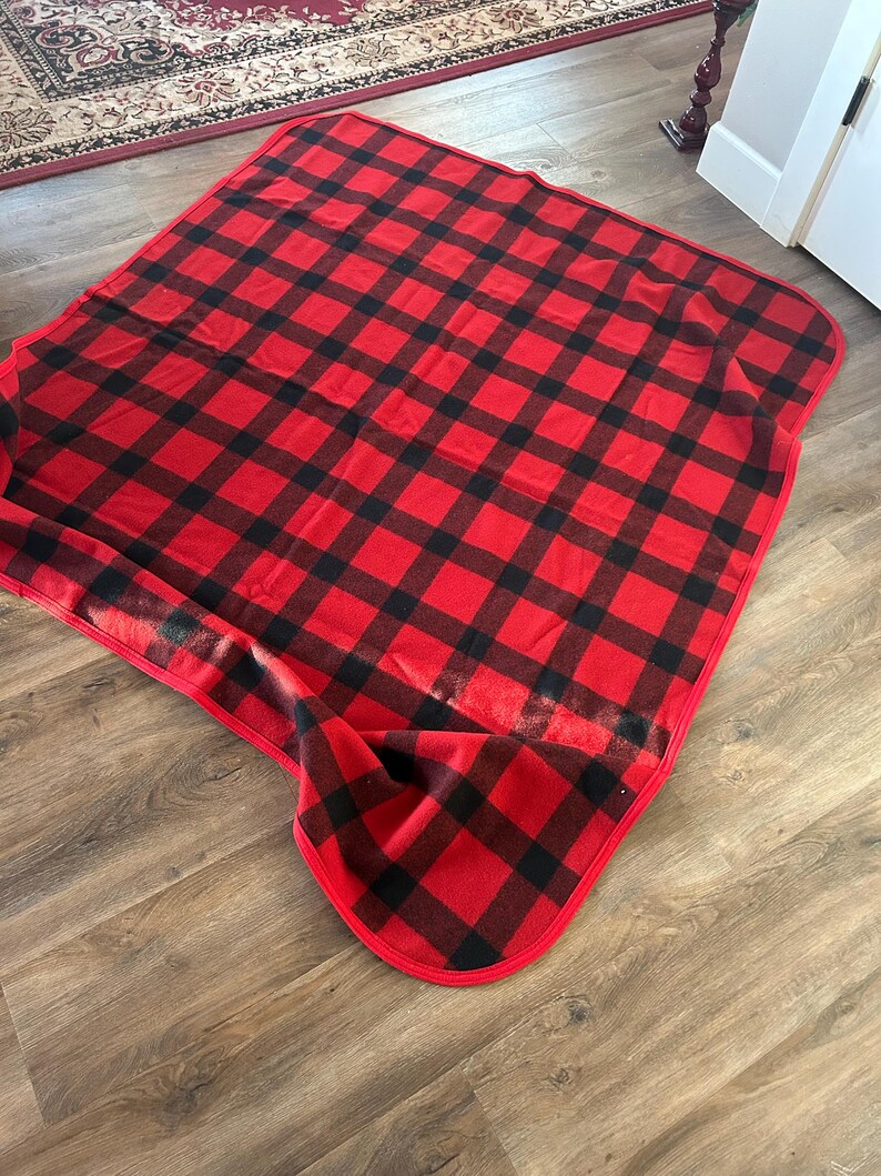 Może przedstawiać: Czerwono-czarny koc w kratę z zaokrągloną krawędzią. Koc jest wykonany z miękkiego materiału i jest złożony na drewnianej podłodze. Wz&oacute;r to klasyczna krata bawola.