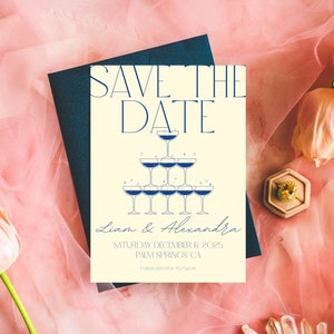 May include: A cream-colored "Save the Date" card with navy blue text and a champagne tower illustration. The card is on a pink fabric background with a navy blue envelope. The card includes the names Liam & Alexandra and the date December 6, 2025.