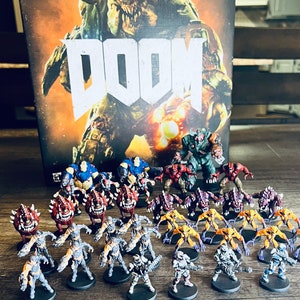May include: A collection of painted miniature figures from the Doom video game. The figures include demons, soldiers, and other characters from the game. The figures are arranged on a wooden table in front of a box with the Doom logo.