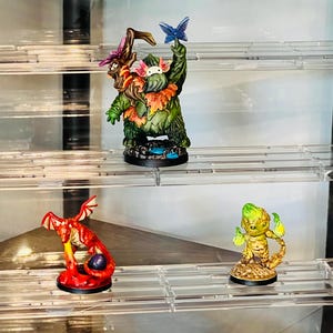 May include: Five miniature figurines displayed on a clear acrylic shelf unit. The figurines are painted in various colors, including green, red, blue, and yellow. The figurines depict a variety of creatures, including a dragon, a fairy, and a tree spirit.
