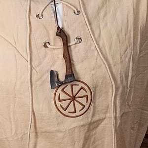 May include: A beige tunic featuring a lace-up neckline with silver-toned metal grommets and long matching cords. An embroidered brown circular symbol with eight radiating spokes is centred on the chest, with a small wooden axe pendant hanging.