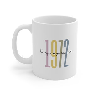 May include: White ceramic mug with the text "leaping since 1972" in colorful vertical lettering.