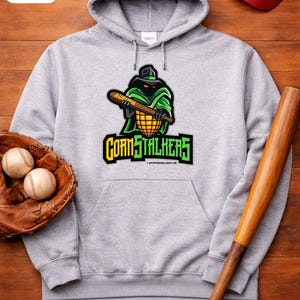 Può includere: Una felpa con cappuccio grigia con un logo "Cornstalkers" raffigurante un personaggio dei cartoni animati che tiene una mazza da baseball. Sono visibili anche un guanto da baseball con due palline, una mazza da baseball e un berretto da baseball rosso.