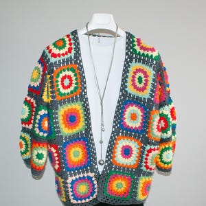 May include: A colorful, handmade crochet cardigan displayed on a white mannequin. The open-front sweater features a patchwork design of vibrant squares in shades of red, orange, yellow, green, blue, and purple, set against a gray background. A silver necklace hangs from the mannequin.