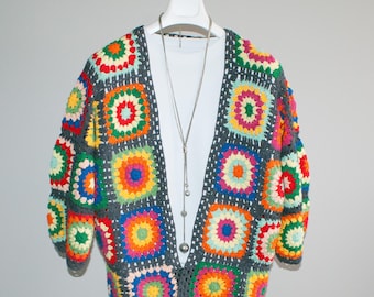 Puede incluir: Un cárdigan de ganchillo hecho a mano y colorido, exhibido en un maniquí blanco. El suéter abierto presenta un diseño de patchwork de cuadrados vibrantes en tonos de rojo, naranja, amarillo, verde, azul y morado, sobre un fondo gris. Un collar plateado cuelga del maniquí.