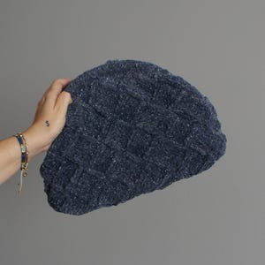 May include: A dark blue knitted beanie hat with a textured, woven pattern. The hat has a soft, plush appearance and is embellished with subtle, sparkling silver threads. The hat is held against a plain grey background.