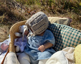 May include: A baby wearing a light brown crocheted hat and a light blue dress is sitting in a wicker basket with a stuffed animal. The basket is surrounded by pillows and blankets.