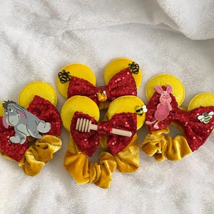 Op de afbeelding: Gele en rode pailletten haaraccessoires met cartoonkarakterversieringen. De collectie omvat scrunchies, strikken en muisvormige oren, met Eeyore, Piglet en honingthema-ontwerpen. De accessoires zijn geplaatst tegen een witte achtergrond.