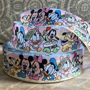 Puede incluir: Un rollo de cinta azul claro y blanco con un patrón repetido de personajes de dibujos animados. Los personajes son Mickey Mouse, Minnie Mouse, Donald Duck, Daisy Duck, Goofy y Pluto. Los personajes llevan todos ropa de bebé y sostienen varios objetos, como un libro, un globo y un sonajero.