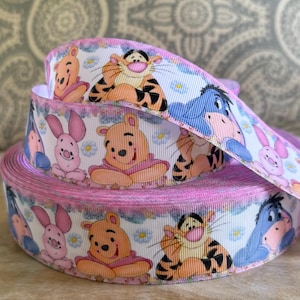 Può includere: Un rotolo di nastro rosa e bianco con personaggi di Winnie the Pooh. Il nastro presenta immagini di Winnie the Pooh, Tigro, Eeyore e Pimpi. Il nastro è decorato con piccoli fiori blu e un bordo rosa.