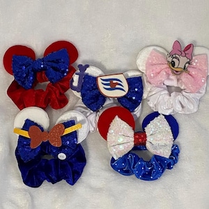May include: A collection of colorful scrunchies with mouse ear designs. The scrunchies feature red, white, and blue colors, with sequined bows and character embellishments. Some have a sequined bow, and others have a character design.
