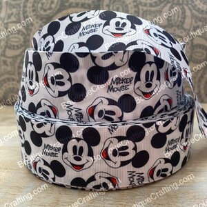 May include: A roll of white ribbon featuring a repeating pattern of Mickey Mouse faces in black and red. The words "Mickey Mouse" are printed in black. The ribbon is likely used for crafting or gift wrapping.