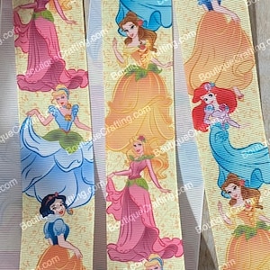 Puede incluir: Una cinta amarilla con un patrón repetido de princesas de Disney. Las princesas son Cenicienta, Ariel, Bella, Blancanieves, Aurora y Rapunzel. Cada princesa lleva su vestido característico y tiene un diseño floral a su alrededor.