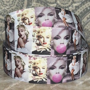 May include: Rolled ribbon with a repeating pattern of Marilyn Monroe in black, white, and pink. The images of Monroe are in black and white. The ribbon features various poses and expressions of the iconic actress.
