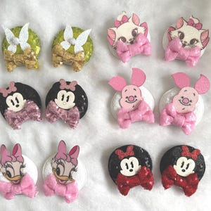 May include: A selection of character hair accessories featuring Minnie Mouse, Marie, Piglet, and Daisy Duck. Each accessory has a character face, a sequined base, and a bow. The colour scheme includes pink, red, black, white, and gold.