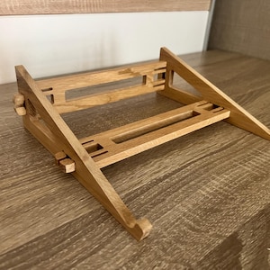 May include: A wooden laptop stand with a natural finish. The stand is angled, with a horizontal base and two angled supports. It features cut-out details and adjustable elements. The stand is designed to elevate a laptop for improved ergonomics.