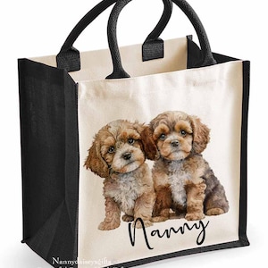 May include: A cream-coloured tote bag with a watercolour illustration of two brown and tan puppies. The word "Nanny" is written in a cursive font below the puppies. The bag has black sides, bottom, and handles.